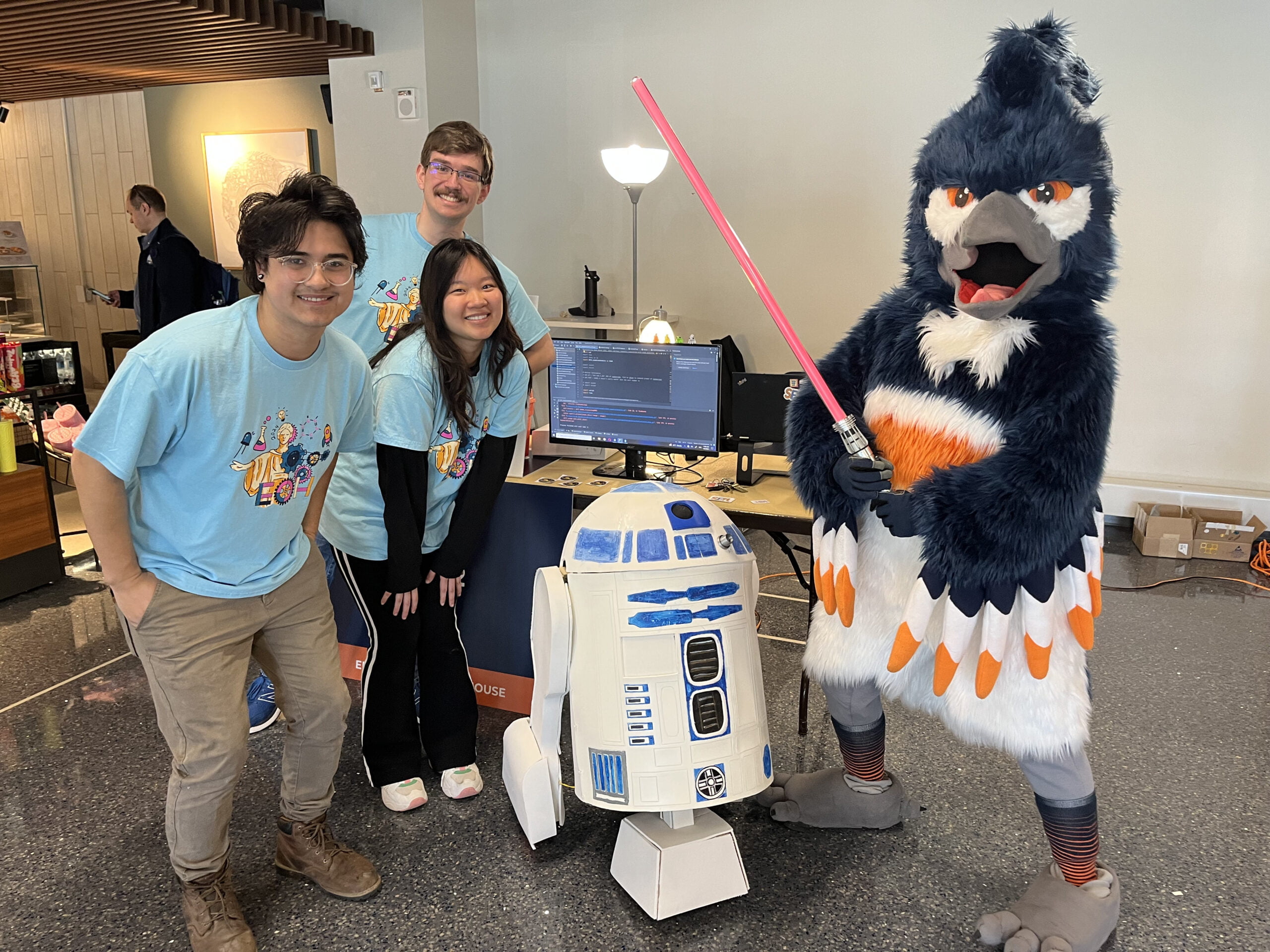 The Belted Kingfisher holding a lightsaber posing next to an R2D2 model and a group of students wearing Engineering Open House shirts