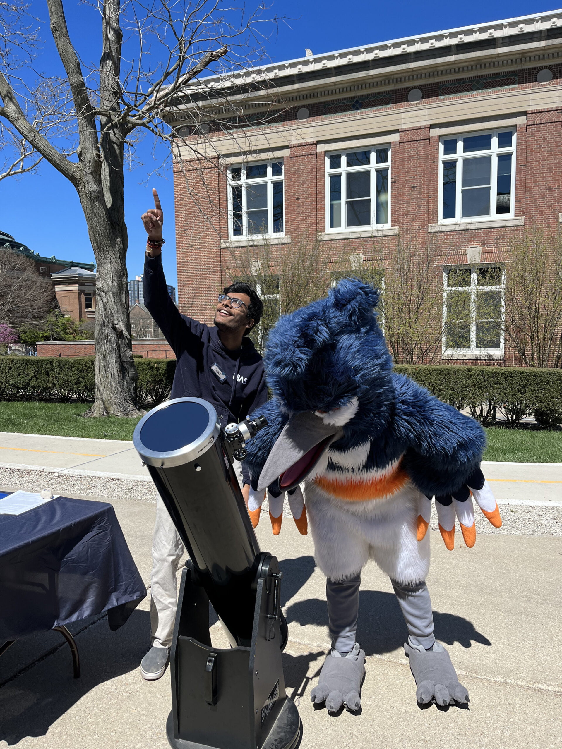 The Belted Kingfisher mascot looking through a telescope while a student points at the sky