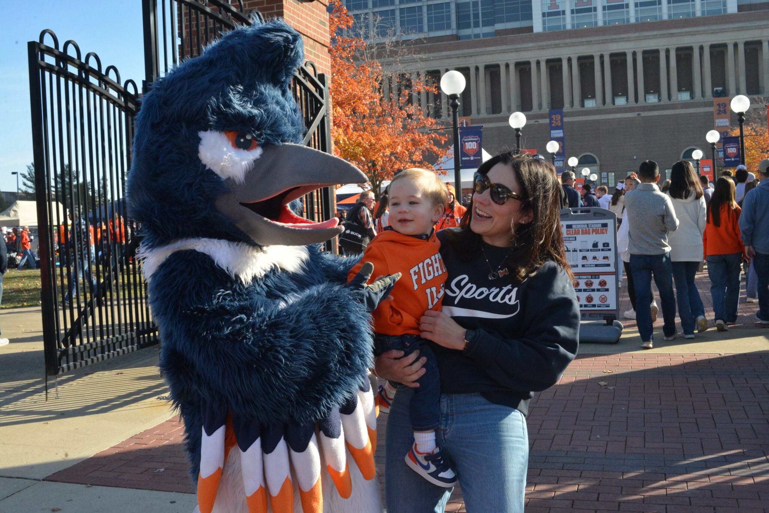 The Belted Kingfisher mascot posing together with an adult and a baby wearing University of Illinois merchandise