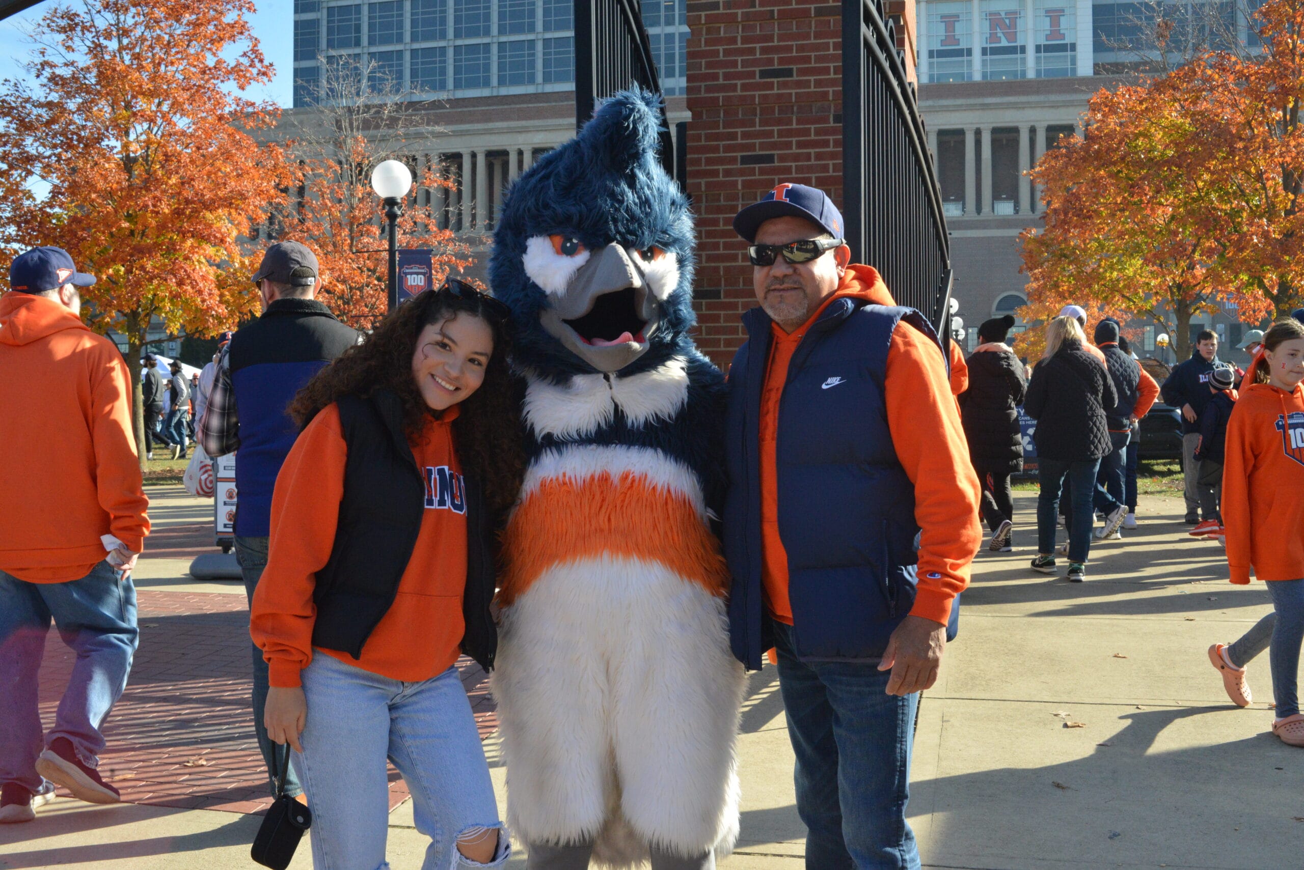 The Belted Kingfisher mascot posing together with two people wearing University of Illinois merchandise