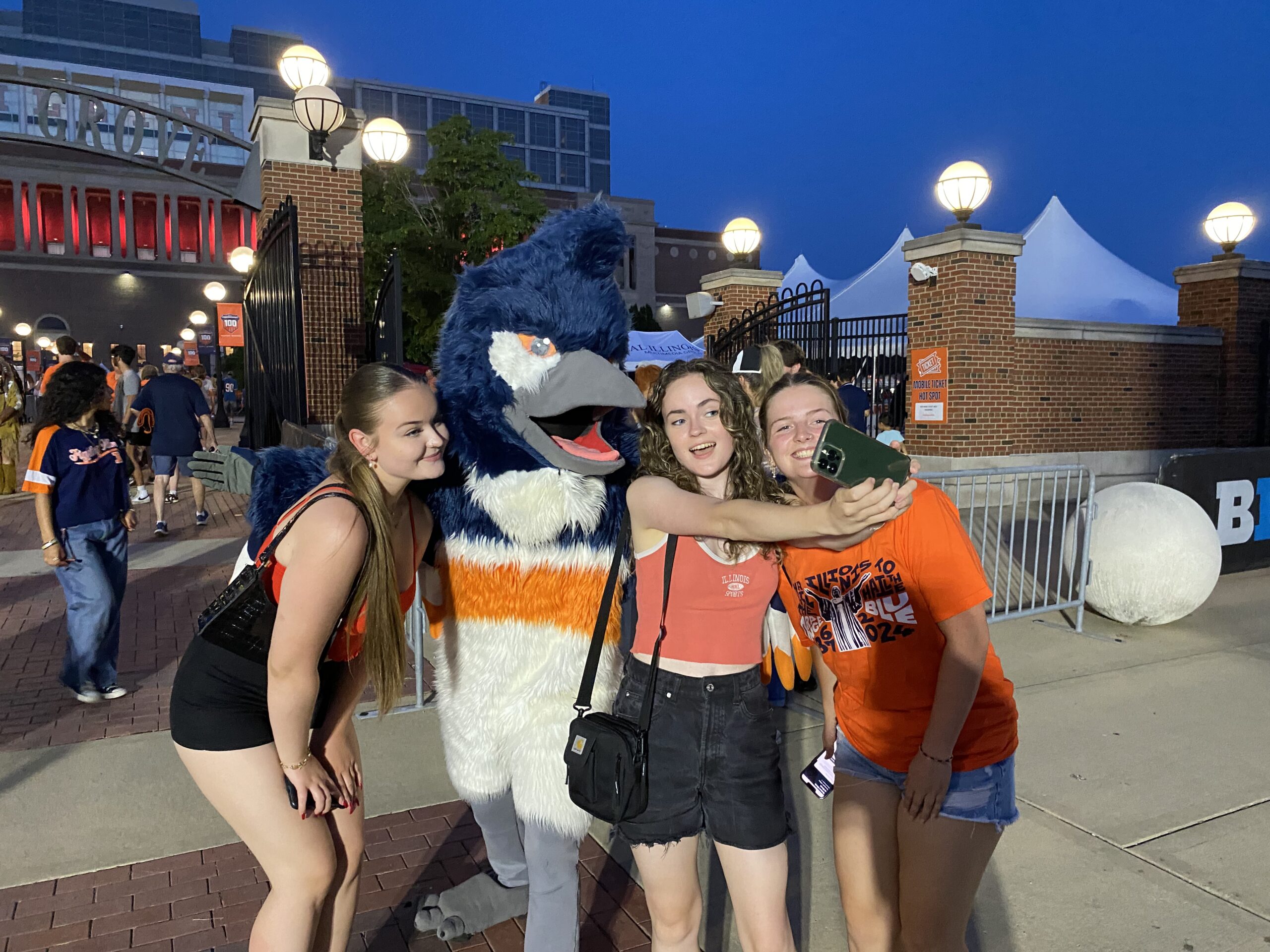 The Belted Kingfisher mascot taking a selfie with 3 students wearing orange and blue