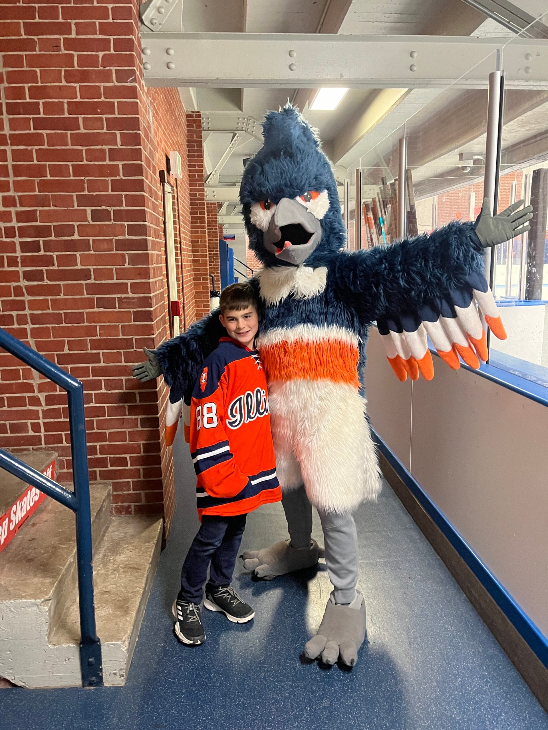 The Belted Kingfisher mascot posing together with a boy wearing an Illinois hockey jersey outside the ice rink