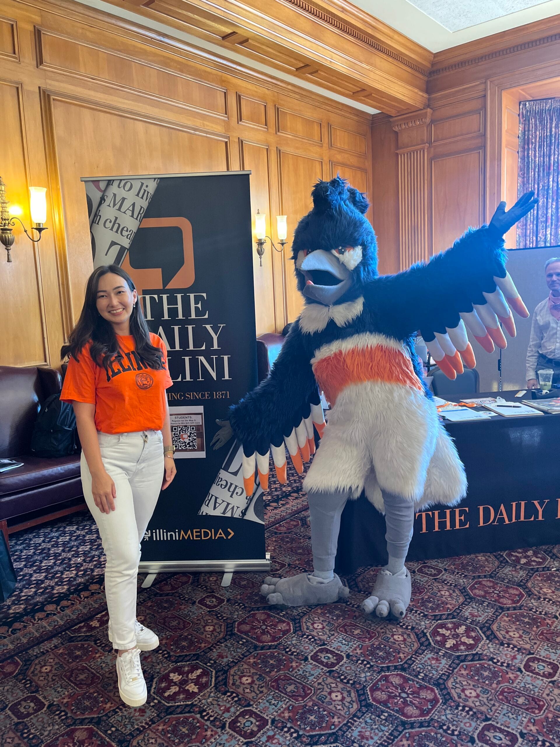 The Belted Kingfisher mascot posing next to an adult at the Daily Illini booth