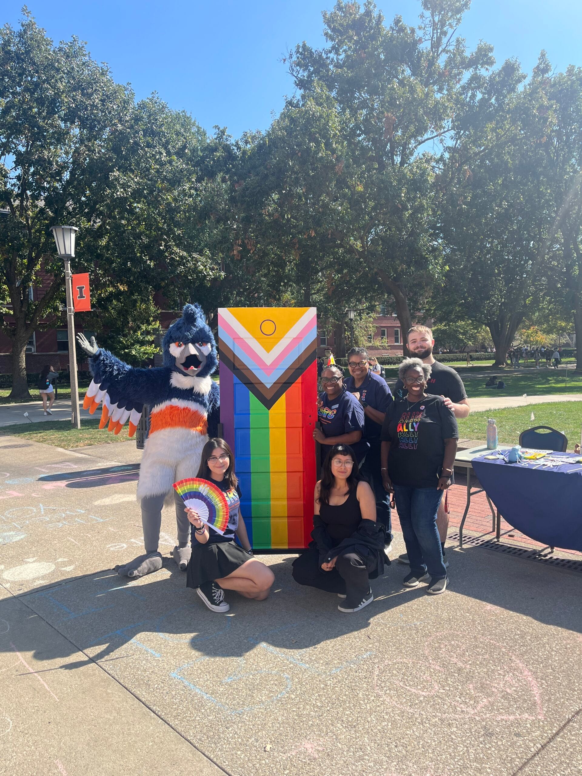 The Belted Kingfisher mascot posing together with 6 adults around an LGBTQIA2S+ flag