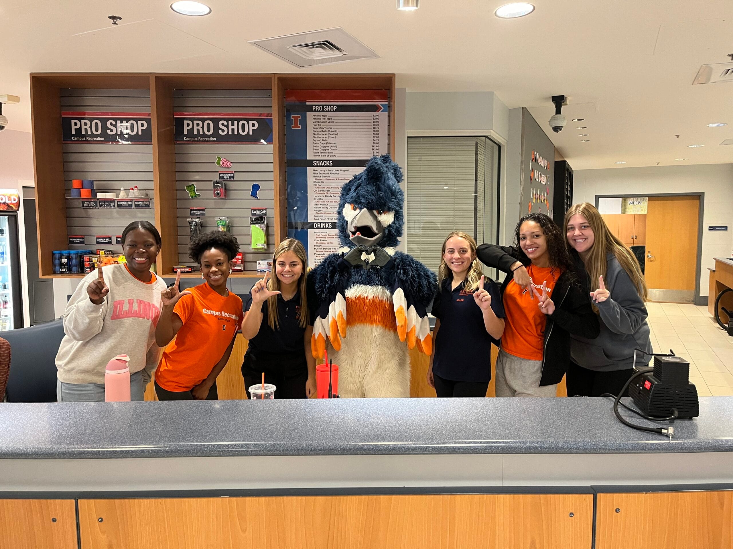 The Belted Kingfisher mascot posing with 6 ARC concierge staff members forming the letters I-L-L-I-N-I with their fingers