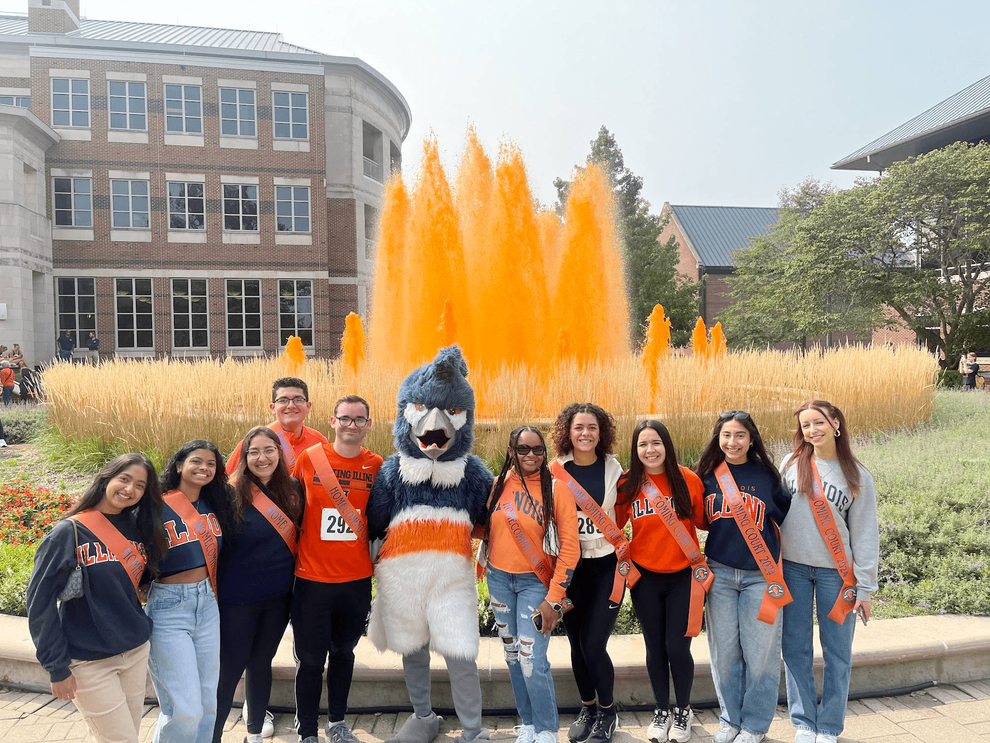 The Belted Kingfisher mascot posing together with10 students from the Homecoming Court in front of an orange fountain