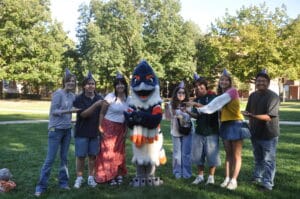 Belted Kingfisher in a party hat holding up a lizard, together with multiple students in party hats