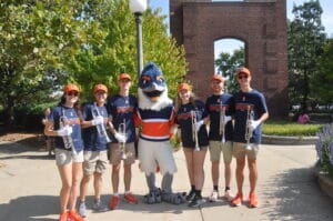 Belted Kingfisher posing with students holding up brass instruments in Homecoming shirts