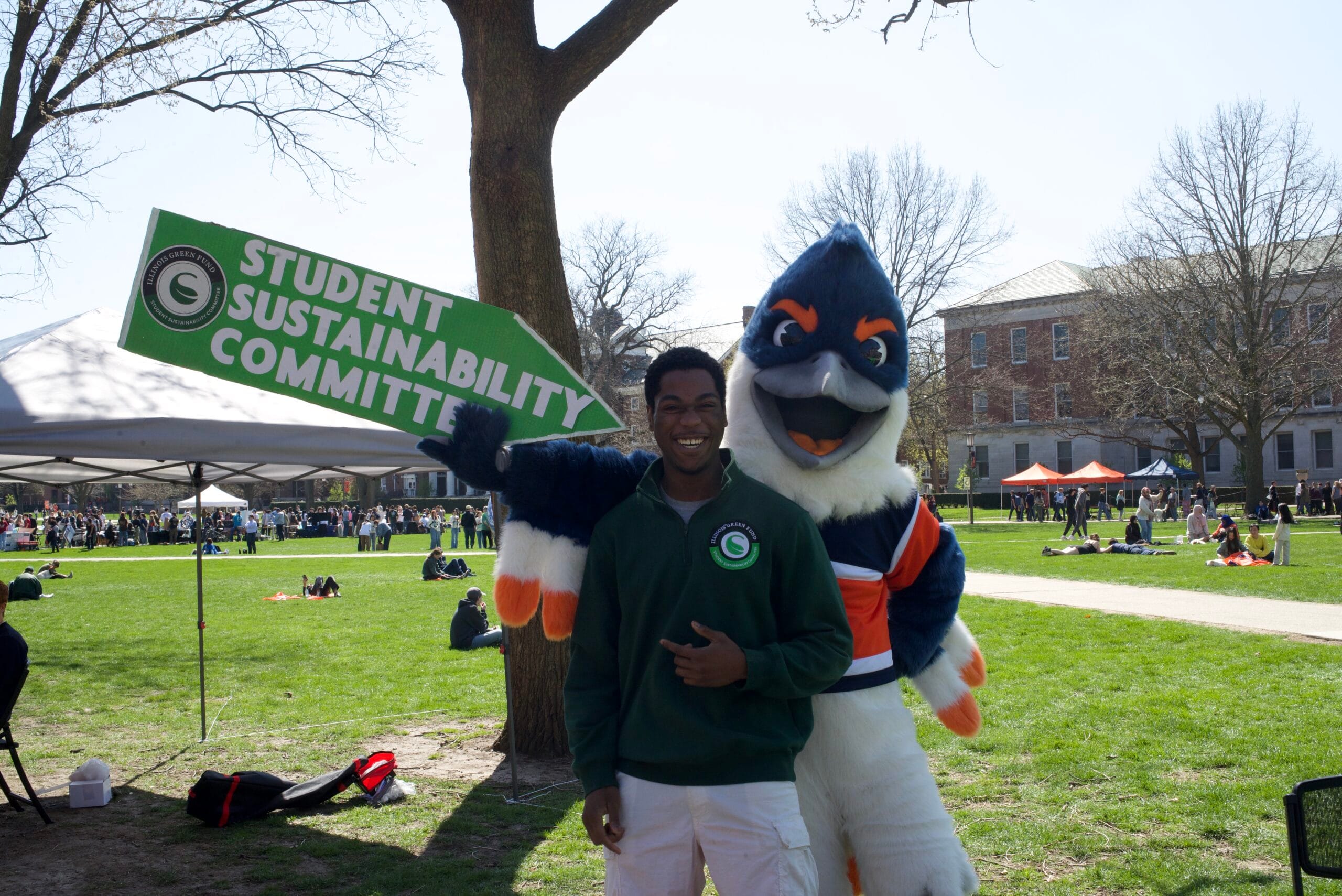 Belted Kingfisher holding up a sign that reads "Student Sustainability Committee", pointing towards a student in a Student Sustainability Committee hoodie