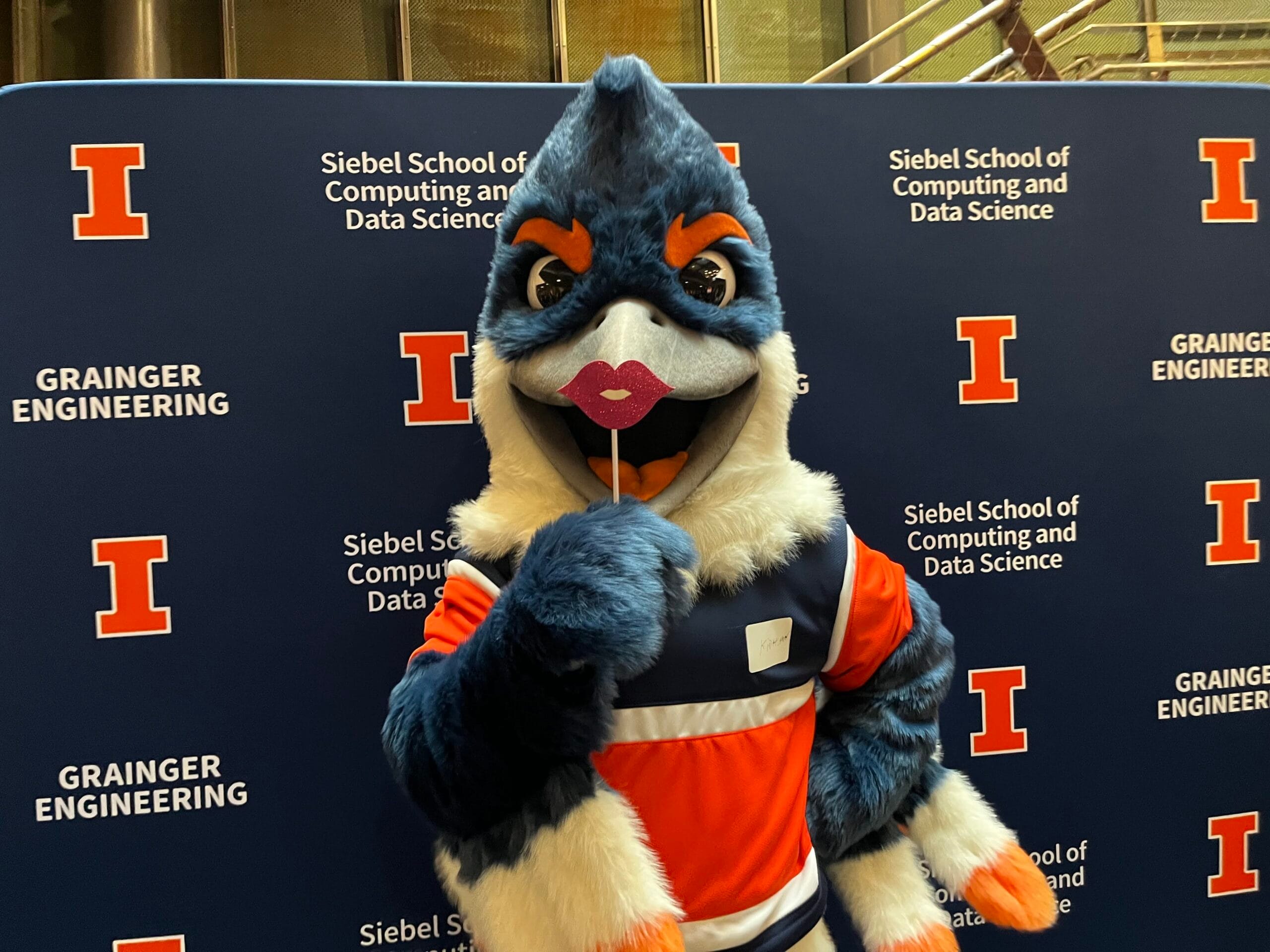 Belted Kingfisher posing in front of a Siebel School of Computing and Data Science backdrop while holding up a photo booth prop shaped like lips