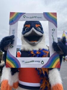 Belted Kingfisher holding up a frame with rainbow corners and the text "You belong here" in different languages along each edge