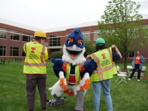 Belted Kingfisher pointing towards the camera while wearing a safety vest, between two people in safety vests with their backs turned towards the camera. The vests read "Prairie Research Institute"