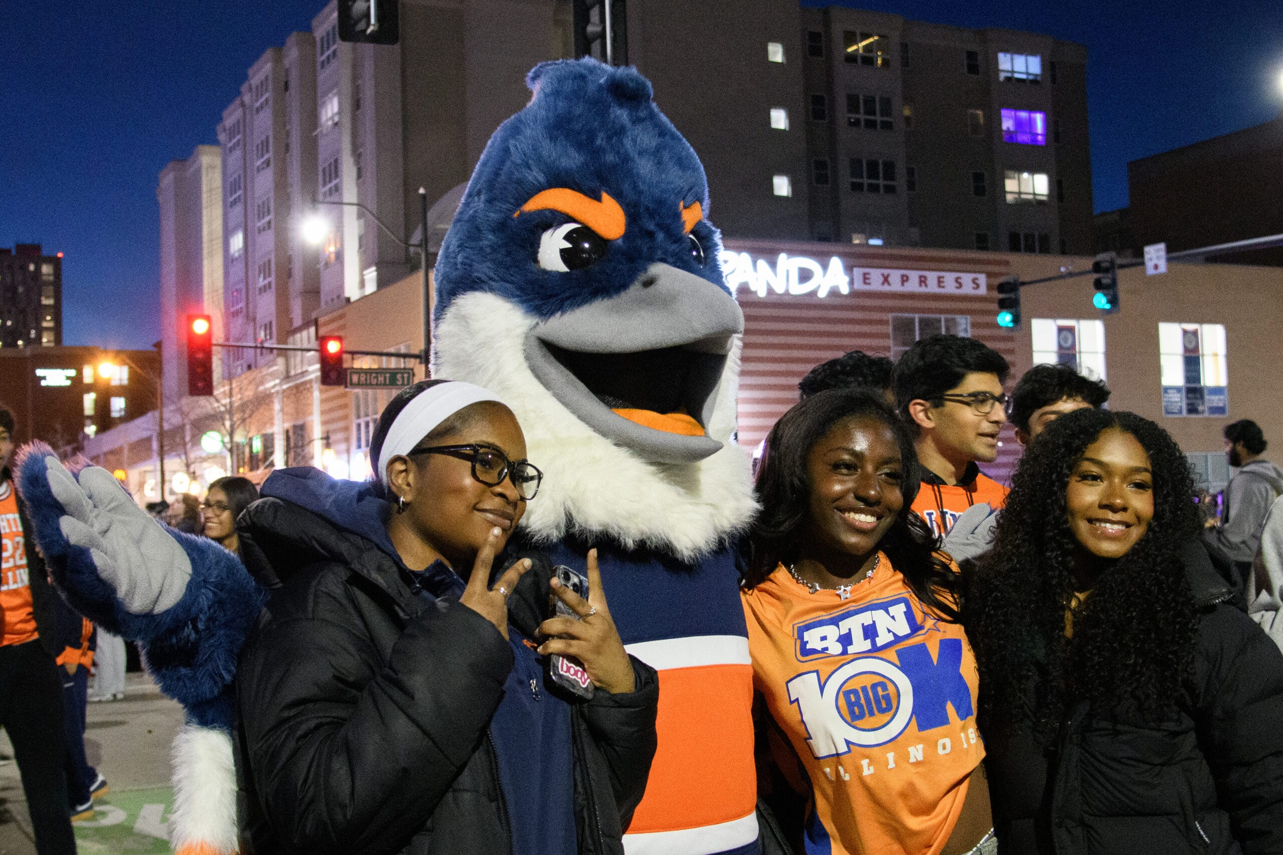 The Belted Kingfisher mascot posing with three University of Illinois students on Green Street. One of the students is wearing a Big Ten Illinois shirt.