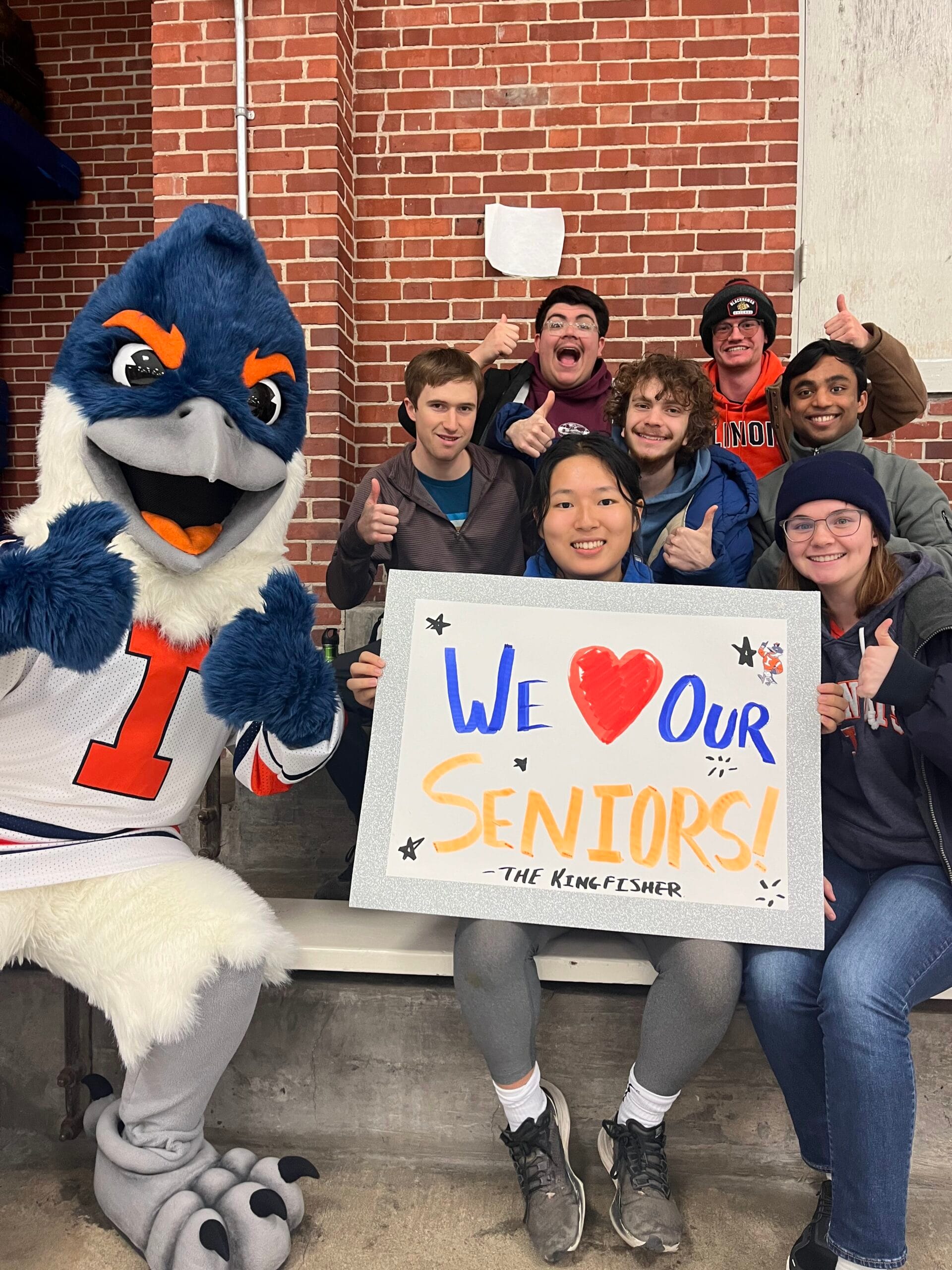The Belted Kingfisher mascot posing with a group of students holding up a sign that reads "We Love Our Seniors!"