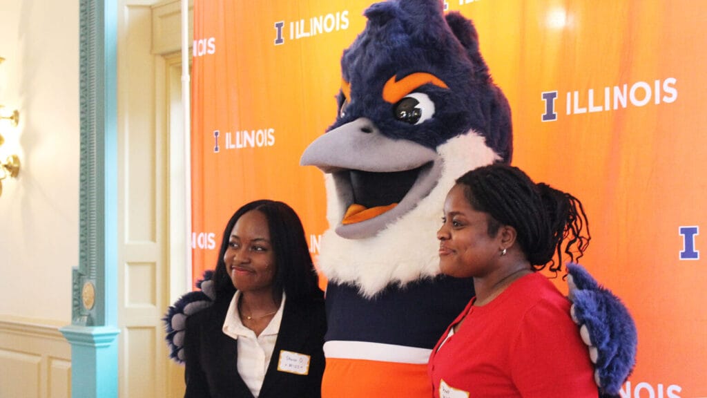 The Belted Kingfisher mascot posing with two students in front of an orange Illinois backdrop