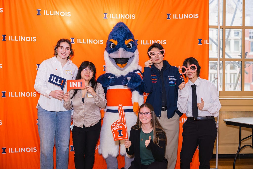 The Belted Kingfisher mascot posing together with students in business attire holding up photo booth props